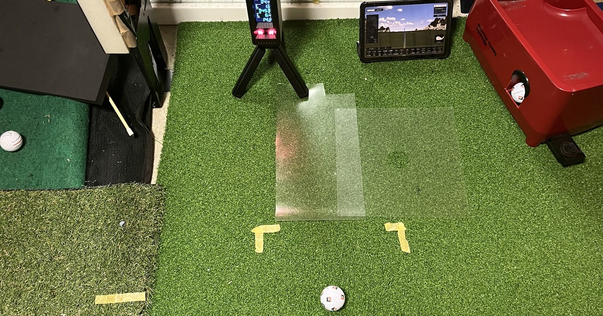 自宅6畳の練習部屋。人工芝の上に弾道測定器が三脚で立ち、その横にタブレット。手前に黄色いL字のマーカーで囲まれたボールが置いてある。